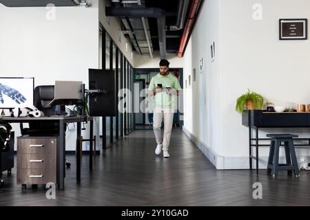 Cammina in ufficio, uomo indiano con smartphone e tazza di caffè, moderno spazio di lavoro Foto Stock