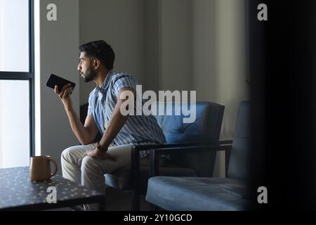 Tenendo in mano lo smartphone, un uomo indiano seduto sulla sedia e guardando fuori dalla finestra in ufficio, spazio fotocopie Foto Stock