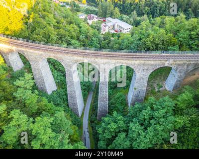 Il ponte ferroviario in pietra di Zampach si estende su una vegetazione lussureggiante, collegando due lati di una valle, con uno sguardo agli edifici vicini che sbirciano attraverso le foglie sotto la luce del sole. Foto Stock