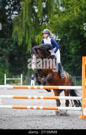 Una giovane donna con una camicia blu e un casco guida un cavallo di baia sopra un ostacolo nella gara di salto ostacoli Foto Stock