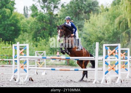 Una giovane donna con una camicia blu e un casco guida un cavallo di baia sopra un ostacolo nella gara di salto ostacoli Foto Stock