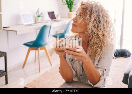 Una donna bionda riccia seduta al tavolo della cucina bevendo un caffè guarda e pensa mentre si rilassa. La donna di mezza età si rilassa prendendo una pausa dal lavoro. Foto Stock