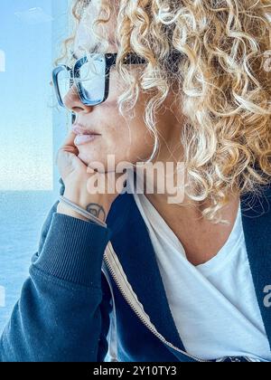 Bella donna con gli occhiali che passa il tempo libero guardando il mare. Si prende cura di se stessa, rilassandosi. Goditi la vista, guarda attraverso la finestra. Bionda di mezza età e donna riccia con un look attento. Mattine tranquille. Foto Stock