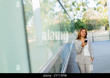 Una donna professionista cammina con facilità, sorridendo mentre si concentra sul telefono, mescolando lo stile di vita moderno con la tranquillità della natura sullo sfondo Foto Stock