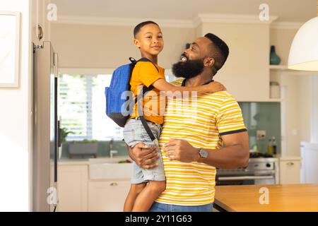 Padre che tiene il figlio con lo zaino, sorridendo insieme in cucina prima della scuola Foto Stock