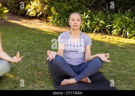 Meditando sul tappetino yoga, donna asiatica anziana che pratica consapevolezza in un giardino all'aperto Foto Stock