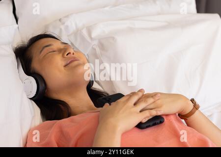 Rilassatevi sul letto, donna asiatica che ascolta musica con le cuffie e tiene in mano lo smartphone Foto Stock