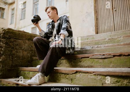 Giovane uomo che tiene la fotocamera seduto sui gradini all'aperto Foto Stock