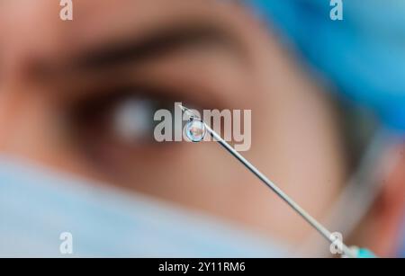 Goccia di medicina sulla punta di ago per iniezione medica. Soluzione iniettabile. Primo piano dell'ago. Medico in maschera medica con siringa preparazione per iniezioni Foto Stock