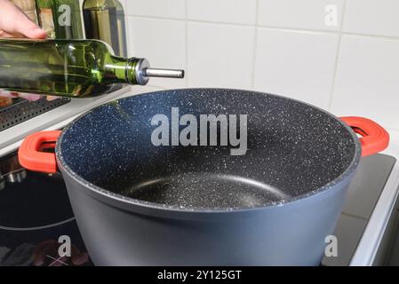 La mano di una donna con una bottiglia di olio. Primo piano. Versare l'olio in una pentola grande sul piano cottura per la cottura Foto Stock