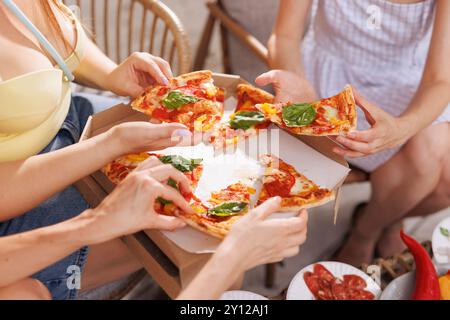 Un gruppo di amici condivide la pizza all'aperto, le mani si avvicinano per le fette guarnite con basilico fresco, mentre una scatola per pizza e piatti di antipasti si siedono sulla schiena Foto Stock