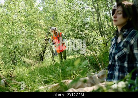 Concentratevi su una donna matura volontaria o bagnino in un giubbotto di sicurezza che cammina lungo il sentiero forestale e guarda a giovani turisti inconsapevoli Foto Stock