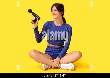 Giovane donna sportiva che utilizza un dispositivo di massaggio a percussione su sfondo giallo Foto Stock