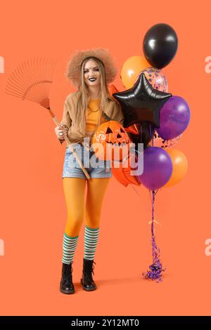 Giovane donna vestita per Halloween con zucca, rastrello e palloncini su sfondo arancione Foto Stock