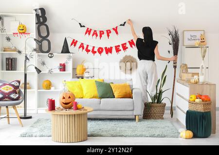 Donna che appende la ghirlanda di Halloween sulla parete luminosa di casa, vista posteriore Foto Stock