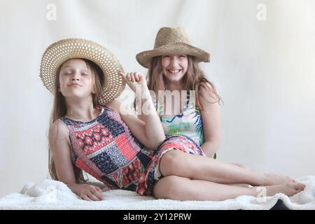 ritratto di due ragazze preadolescenti felici in abiti da sole e cappelli di paglia seduti su una coperta bianca che ridono e si divertono sullo sfondo chiaro Foto Stock