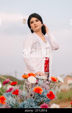 Il fascino di una giovane donna asiatica che regge candidamente un panino che indossa moderni abiti tradizionali giavanesi, un kebaya nuziale in velluto bianco in un'altra parte Foto Stock