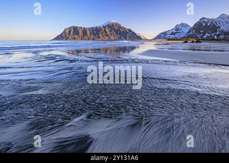 Corso fluviale sulla spiaggia di fronte a montagne innevate, luce del mattino, inverno, Flakstadoya, Lofoten, Norvegia, Europa Foto Stock