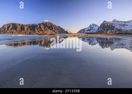 Montagne innevate che si riflettono in acqua sulla spiaggia, mare, luce mattutina, inverno, Flakstadoya, Lofoten, Norvegia, Europa Foto Stock