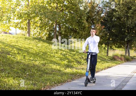 Gioioso e uomo informale equitazione scooter elettrico nel parco della città e parlando al telefono Foto Stock