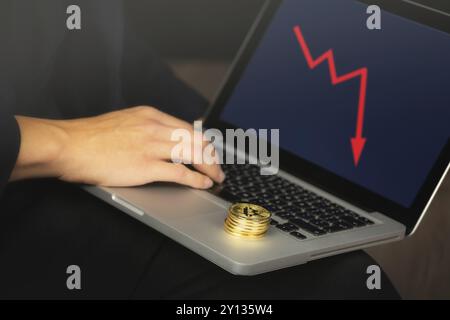 Uomo d'affari che lavora su un laptop con una pila di monete bitcoin dorate. Grafico rosso, concetto di crash dei prezzi Foto Stock