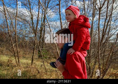 Un ragazzo fornisce un passaggio alla sorellina, che indossa un vestito rosso brillante, mentre si godono una calda passeggiata pomeridiana in una scenografica area boschiva fille Foto Stock