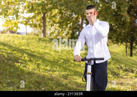 Gioioso e uomo informale equitazione scooter elettrico nel parco della città e parlando al telefono Foto Stock