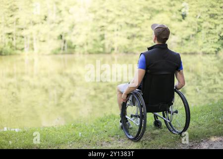 Uomo felice e giovane disabile o handicappato seduto su una sedia a rotelle che guarda il bellissimo lago in natura Foto Stock