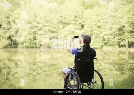 Uomo felice e giovane disabile o handicappato seduto su una sedia a rotelle in natura. Fotografare paesaggi con lo smartphone Foto Stock