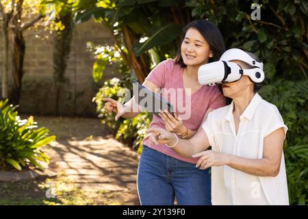 Utilizzando il visore VR, la nonna asiatica esplora la realtà virtuale con assistenza all'aperto Foto Stock