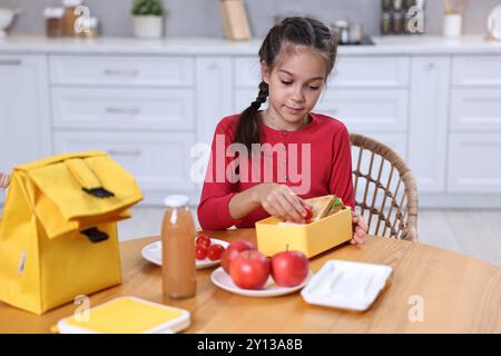 Una ragazza carina che mette i pomodori nel cestino da pranzo al tavolo di legno in cucina. Prepararsi per la scuola Foto Stock