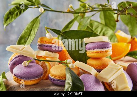 Vivaci macaron gialli e viola visualizzati con fette arancioni, foglie verdi e pezzi di cioccolato bianco su una tavola rustica di legno. Foto Stock