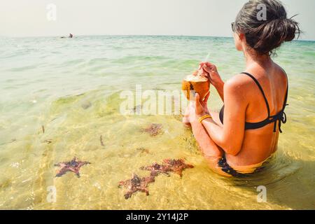 Una donna felice in bikini sulla spiaggia di stelle marine beve una bevanda fresca al latte di cocco sulla spiaggia di pesce stellato godendoti la famosa isola tropicale di Koh Rong, Cambogia. Foto Stock