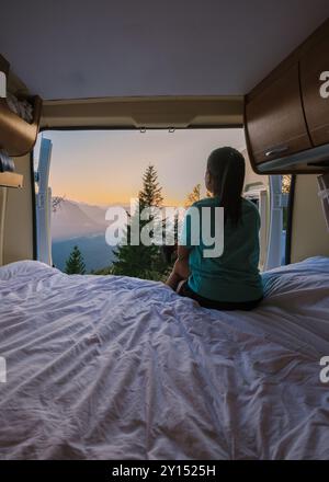 Mentre l'alba si infrangeva su Hallstatt, un viaggiatore siede tranquillamente su un letto all'interno di un camper, osservando lo splendido paesaggio di montagna. Il sole che sorge getta un caldo bagliore sul tranquillo paesaggio. Foto Stock