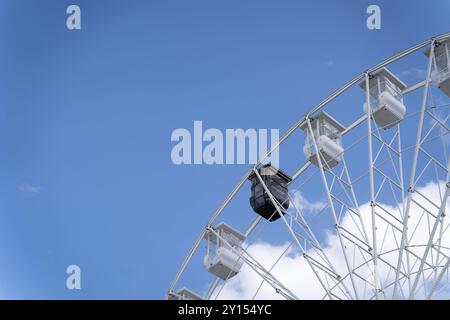 Vista parziale di una ruota panoramica con cabine bianche e solo una nera che crea contrasto, in un parco divertimenti contro un cielo azzurro limpido con alcune nuvole. Foto Stock