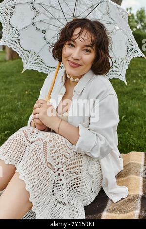 Una giovane donna allegra si rilassa su una coperta, assaporando un picnic estivo con un delicato ombrello. Foto Stock