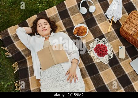 Una giovane donna riposa tranquillamente su una coperta da picnic, circondata da cibo delizioso sotto il caldo sole estivo. Foto Stock