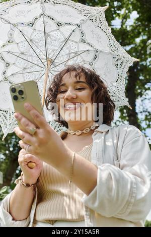 Una giovane donna allegra, vestita di bianco, gode di una giornata di sole in un lussureggiante parco verde mentre controlla il telefono. Foto Stock