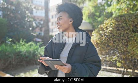 La donna afroamericana con i capelli ricci ama la musica tramite auricolari wireless mentre tiene in mano un tablet all'aperto. Foto Stock