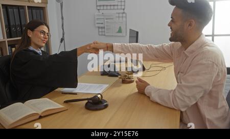 Giudice donna che stringe la mano a un cliente uomo in ufficio, discutendo questioni legali con documenti, martelletto e scale di giustizia sulla scrivania di legno indo Foto Stock
