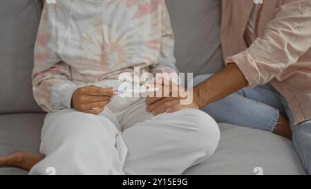 Coppia seduta vicino sul divano in casa con donna che tiene il test di gravidanza e uomo che le tocca delicatamente la mano con un gesto di supporto Foto Stock