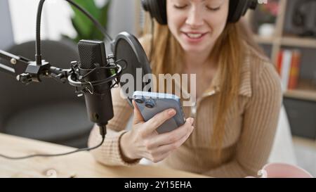 Una giovane donna sorridente con i capelli biondi registra un podcast in uno studio, tenendo in mano uno smartphone. Foto Stock