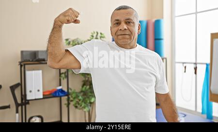 Un uomo ispanico fiducioso che flessa i muscoli in una sala di terapia di un centro di riabilitazione, promuovendo il benessere e la salute. Foto Stock