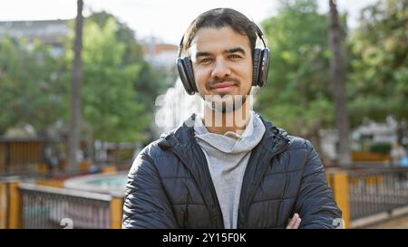 Bel giovane ispanico con la barba che indossa cuffie e giacca in posa all'aperto in un verde parco urbano. Foto Stock