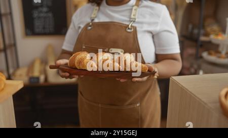 La giovane donna afroamericana in una panetteria tiene in mano un vassoio di croissant, vestito con un grembiule sopra una camicia bianca, scena interna con pas appena sfornati Foto Stock