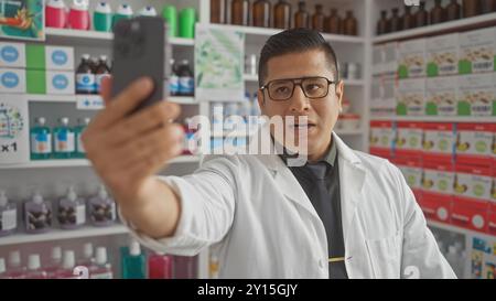 Uomo ispanico che fa un selfie in una farmacia con un camice da laboratorio con scaffali di prodotti sullo sfondo Foto Stock