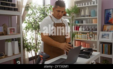 Giovane in grembiule che lavora in un negozio arredato con gusto, organizza prodotti in una stanza piena di vari oggetti decorativi e verde lussureggiante Foto Stock