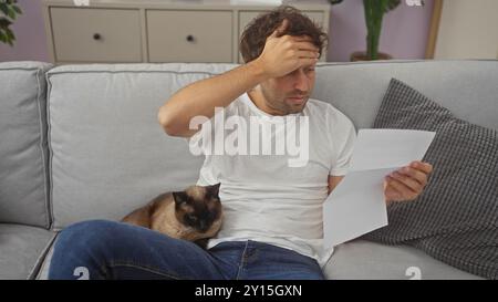 Un giovane preoccupato si siede su un divano grigio leggendo documenti con un gatto siamese accanto a lui in un salotto moderno Foto Stock