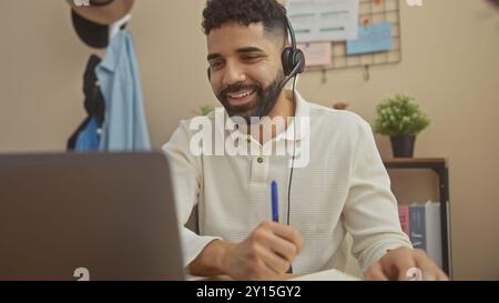 Un giovane allegro con un auricolare impegnato in una conversazione remota nel suo moderno appartamento, penna in mano, lavoro. Foto Stock
