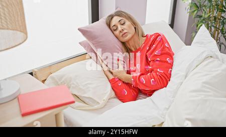 Una donna in un pajama rosso riposa in una camera da letto, gli occhi chiusi, abbracciando un cuscino rosa, evocando relax e comfort. Foto Stock